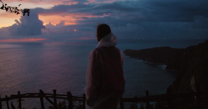Woman in raincoat looks at the sunset over the ocean on Nusa Penida Bali