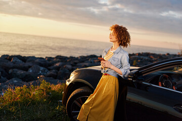 Fototapeta premium Smiling woman with curly hair beside a car on a beach at sunset. Casual fashion, relaxed lifestyle, outdoor vibe, coastal scenery, warm light enhances confident pose.