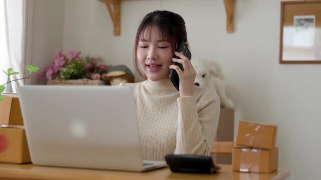 Young woman running an online shop from a home office, calling on a smartphone while managing orders on a laptop with shipping boxes ready for packaging and dispatch.