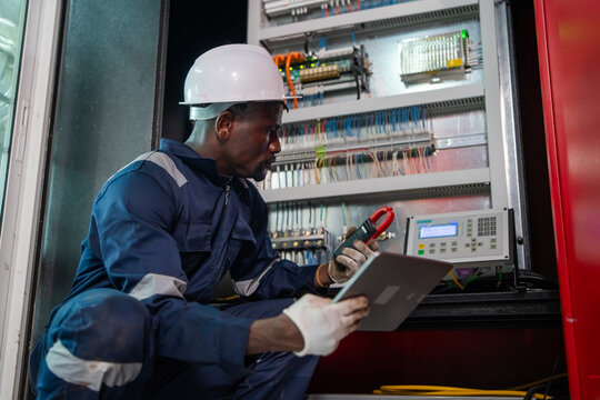 Electrician in blue uniform and white hard hat examines electrical panel. He holds tablet and red clamp meter while reviewing wiring. Panel features relays, wires, and digital display