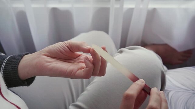 A woman sits by a window filing her nails during a quiet at-home manicure, emphasizing personal grooming, self-care, relaxation and everyday beauty maintenance