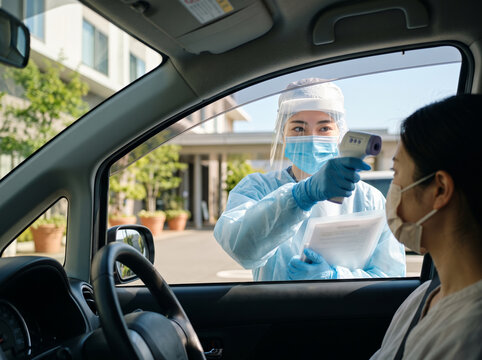 駐車場の車内にいる発熱患者に対して非接触体温計で検温を行なう防護服を着用した医療スタッフの女性