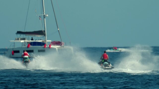 People riding jet skis creating splashes near a catamaran in the ocean, showing water sports and summer leisure activities, Antibes, France
