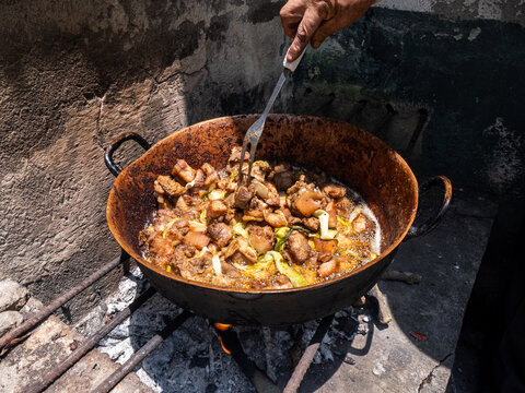 Hand stirs meat in a rusty wok over open flame. Fried chunks sizzle in oil, garlic, and greens. Dusty stone wall frames the rustic cooking scene. Hot air rises from the fire beneath the wok, Fritada