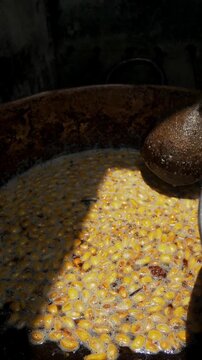 Hand stirs meat in a rusty wok over open flame. Fried chunks sizzle in oil, garlic, and greens. Dusty stone wall frames the rustic cooking scene. Hot air rises from the fire beneath the wok, Fritada