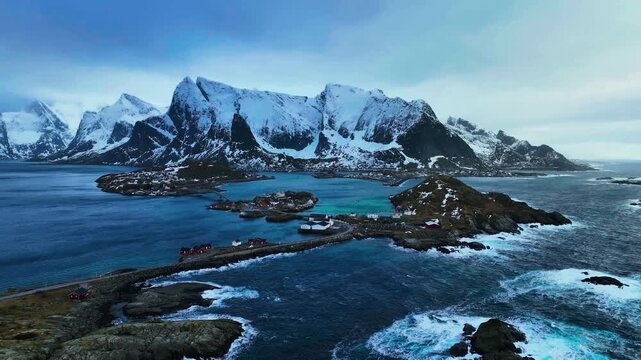 A sweeping, high-altitude drone sequence capturing the iconic fishing village of Reine nestled beneath the dramatic, snow-dusted peaks of Reinebringen.