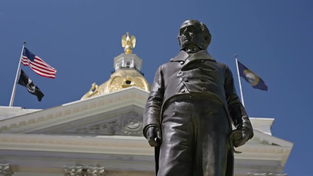 Cinematic orbiting shot looking upward around the Daniel Webster statue on the grounds of the New Hampshire State House in Concord New Hampshire during a bright summer day.