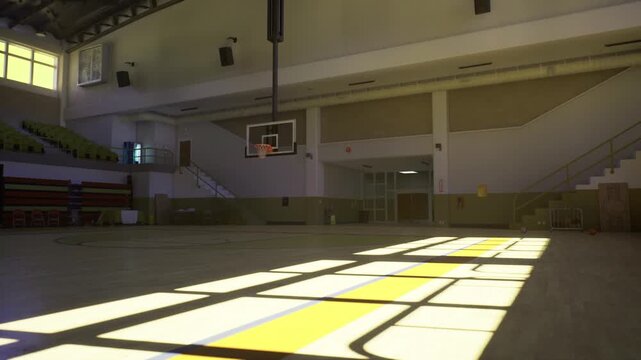 Sunlit empty indoor basketball court with golden hour beams across wooden floor, long shadows from windows, distant hoop and empty bleachers, cinematic