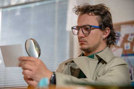 Young adult man in glasses and a trench coat examining a card with a magnifying mirror like a focused detective at work.