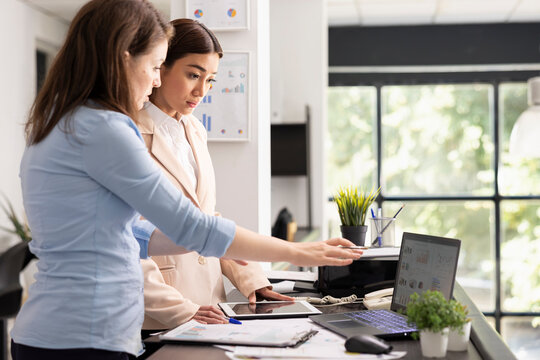 Professional women stand beside desk analyzing financial charts on laptop and communicating about company updates while serious female employees work together to support effective planning.
