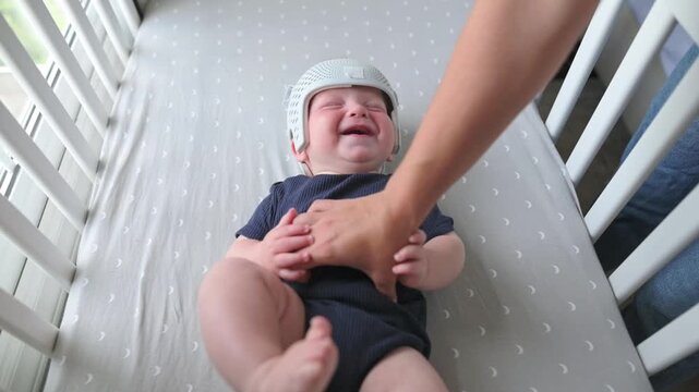 Baby in helmet smiles as caregiver interacts playfully, highlighting a nurturing moment in a well-lit nursery with soft bedding