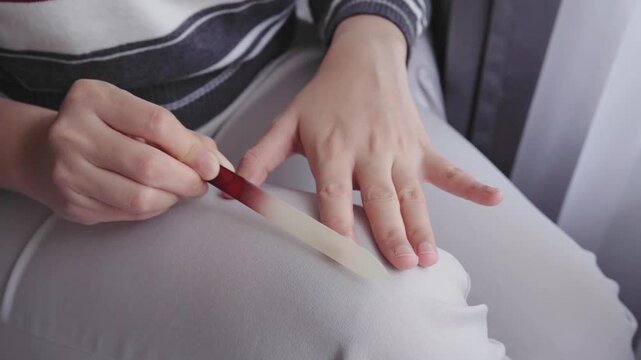 A woman sits by a window filing her nails during a quiet at-home manicure, emphasizing personal grooming, self-care, relaxation and everyday beauty maintenance