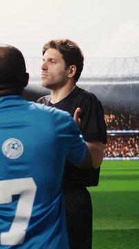 Vertical frame: Soccer referee in black showing yellow card to protesting player on pitch, teammates confronting match official and complaining about decision, disciplinary action moment.