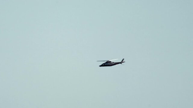 Dark twin-engine helicopter flying horizontally against a clear, light blue sky, representing air travel and transportation