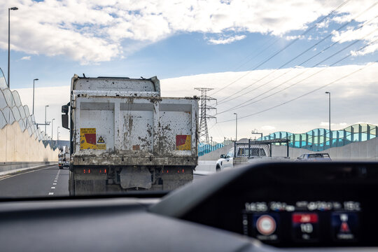 A driver's perspective inside a car, following a large, mud-splattered tipper truck on a busy highway in Melbourne, Australia. Driving safety, heavy vehicle blind spots, industrial transport