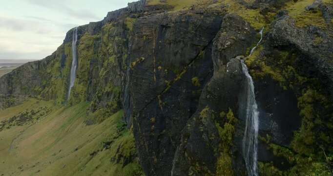 Seljalandsfoss cascading from towering basalt cliffs along Seljalandsa River in Rangarthing eystra Iceland, revealing volcanic escarpments, moss-covered rock faces and adjacent waterfalls drone reveal