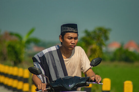 Young Indonesian Muslim guy riding a motorcycle on a village road, heading for Friday prayer or Eid celebration during his sunny daylight journey.