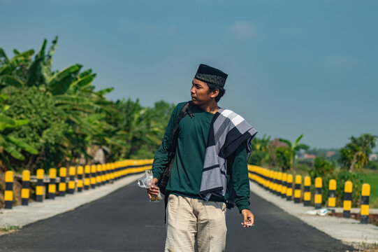Asian Muslim man looking sideways while walking on a rural empty highway during the day, wearing a traditional peci hat and casual attire.
