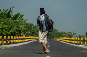 Fototapeta premium Back view of Muslim man with backpack standing on rural road, preparing for Eid homecoming or traditional mudik journey.