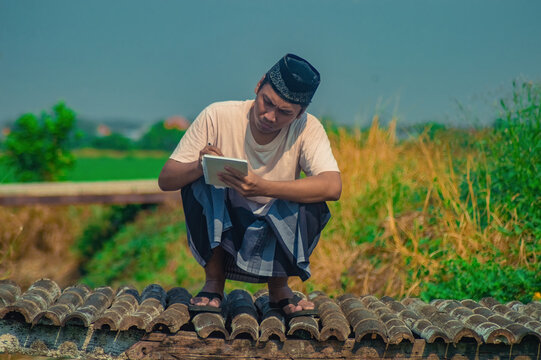 Asian Muslim man wearing a traditional sarong and peci hat squatting on a rural bridge while writing in a notebook with a pen under a bright blue sky.