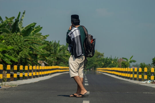 Back view of Muslim man with backpack standing on rural road, preparing for Eid homecoming or traditional mudik journey.