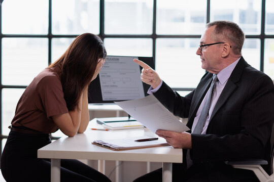 Stressed young woman covering her face, receiving an angry reprimand from her male supervisor during a difficult one on one meeting in an office setting