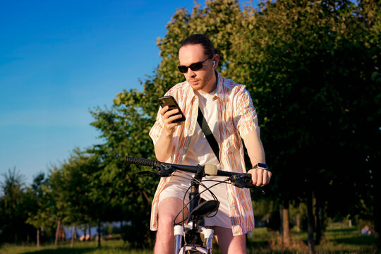 Casual man with a ponytail checks his smartphone while riding a bicycle through a scenic urban park on a bright and sunny summer morning in the city center