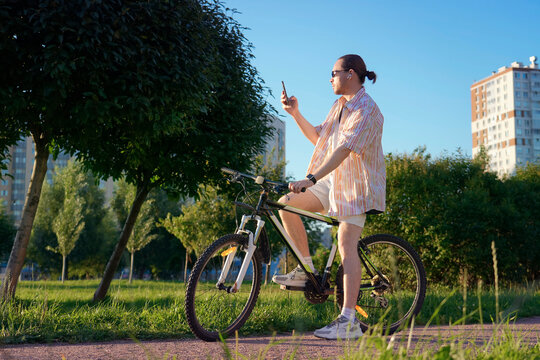 Man sits on a mountain bike in a public park during a sunny summer morning and uses a mobile phone to stay connected while enjoying his ride through the green city