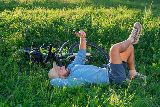 Smiling guy rests on the meadow during a summer trip and uses his phone to communicate while lying on the grass next to his bicycle