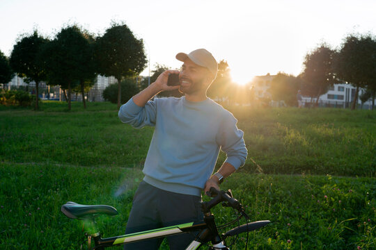 Cheerful guy enjoys a peaceful summer morning in a city park while talking on a smartphone and standing next to his bicycle under the warm sunrise light