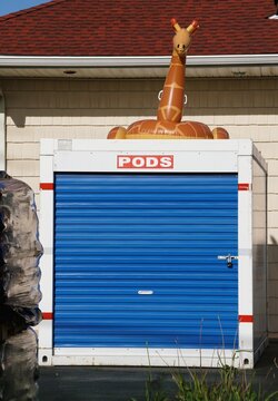 Sussex County, Delaware, U.S.A - July 1, 2026 - A white PODS storage container topped with a large inflatable giraffe float beside a beige building.