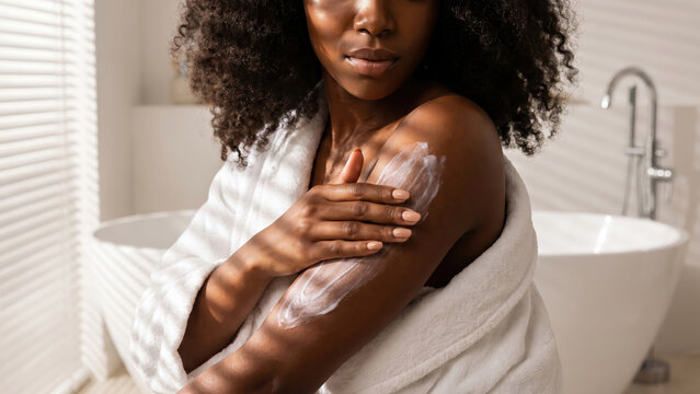 Black woman applying moisturizing body lotion for healthy skin in a bright bathroom.