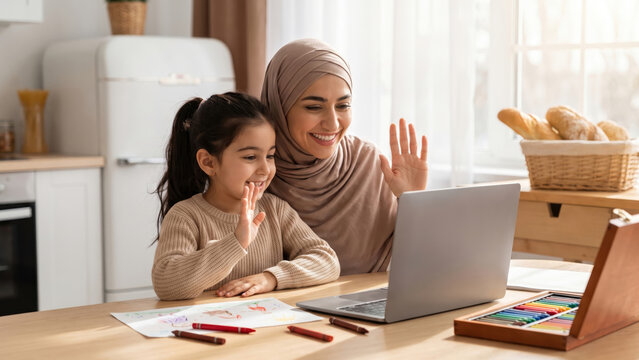 Happy Muslim mother and daughter waving during video call on laptop at home.