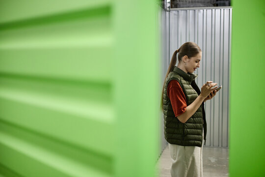 Young adult woman using smartphone beside self storage unit, managing rental access and inventory. Ideal for moving services, personal storage, mobile booking, urban lifestyle marketing