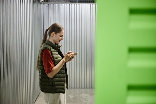 Young adult Caucasian woman standing in self storage corridor, using smartphone while managing personal unit access and inventory. Useful for moving, rental, decluttering marketing