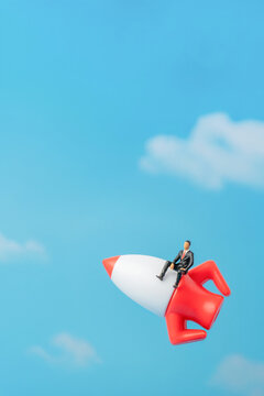 Businessman figurine sitting on a rocket ship flying upwards through a blue sky with clouds. Startup launch, career growth, acceleration, ambition concept.