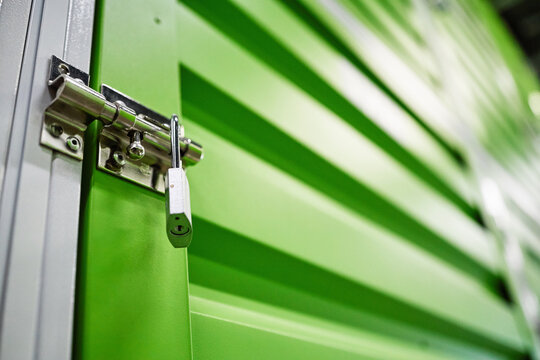 Close up of storage unit door showing locked latch and padlock, securing individual belongings in self storage facility. Supporting rental, moving, security, decluttering marketing