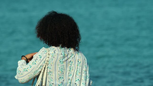 Woman with afro hairstyle stands alone, looking at the vibrant turquoise-blue ocean. Solitude and freedom concept