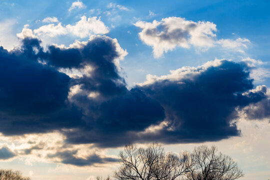 Dramatic contrast of dark stormy clouds rolling across a bright blue sky above the silhouette of bare winter trees, capturing a moody transition between weather patterns at dusk