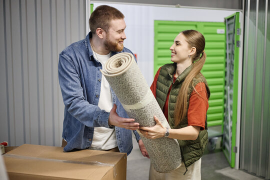 Young adult couple carrying rolled carpet into self storage unit, organizing personal belongings during move. Ideal for relocation, decluttering, rental storage marketing campaigns