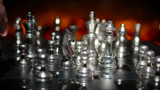 Silver chess pieces being set up or rearranged on a board with dramatic warm orange fire-like lighting in the background.
