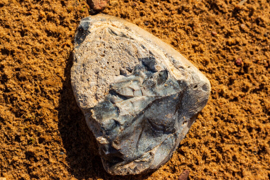 Raw flint stone with sharp edges rests on coarse golden sand at a gravel quarry, showcasing natural mineral textures and geological formation details under bright sunlight