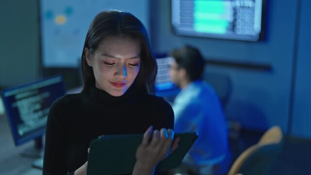 Young Asian woman in black turtleneck working on digital tablet, male developer at laptop in background, tech team in dark blue office.