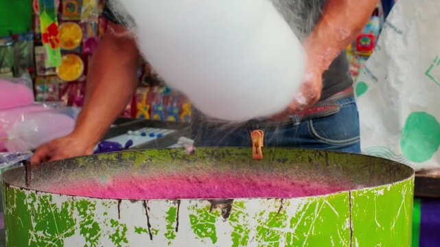 Fresh cotton candy spinning at colorful carnival food stall close up as vendor pours more crystals into drum
