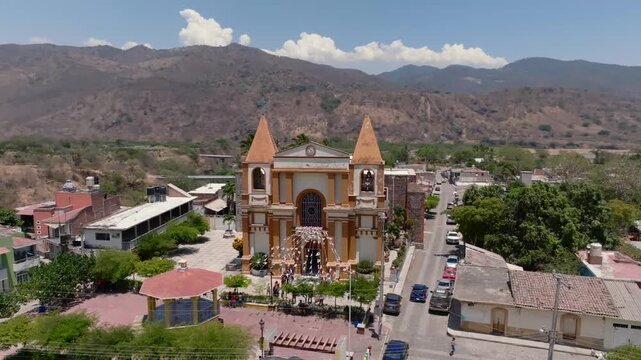Aerial orbit around Platanar town center overlooking the Parroquia de la Virgen de Platanar church facade, central plaza and surrounding mountains in Jalisco, Mexico on a sunny day