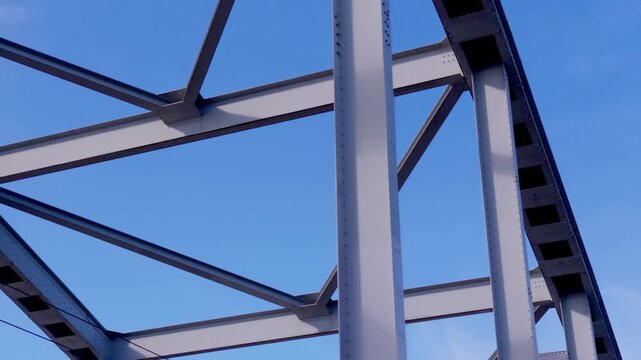 Slow pan reveals intricate steel framework and strong support beams of John Frost Bridge in Arnhem, Netherlands, set against clear blue summer sky.