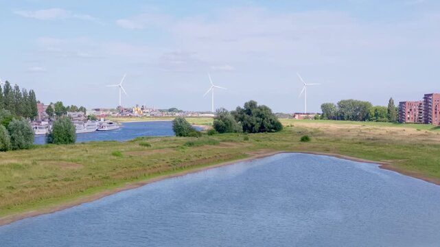 Wide elevated shot capturing the Rhine River, adjacent pond, green landscape, and modern wind turbines symbolizing renewable energy development on the outskirts of Arnhem city.