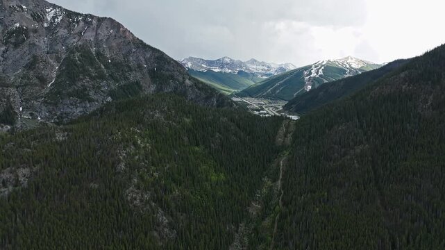 Alpine valley leading toward snowy peaks beyond Officer's Gulch, drone dolly to distant community and highway