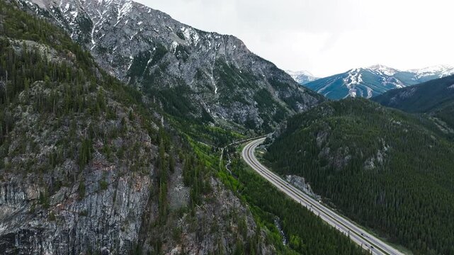 Snowy alpine peaks above Officer's Gulch highway and evergreen forest, drone