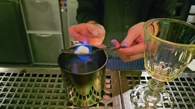 Close view of bartender preparing absinthe at a cocktail bar, with blue fire, sugar cube, slotted spoon, metal cup, glass goblet, stainless counter, and nightlife ambiance.